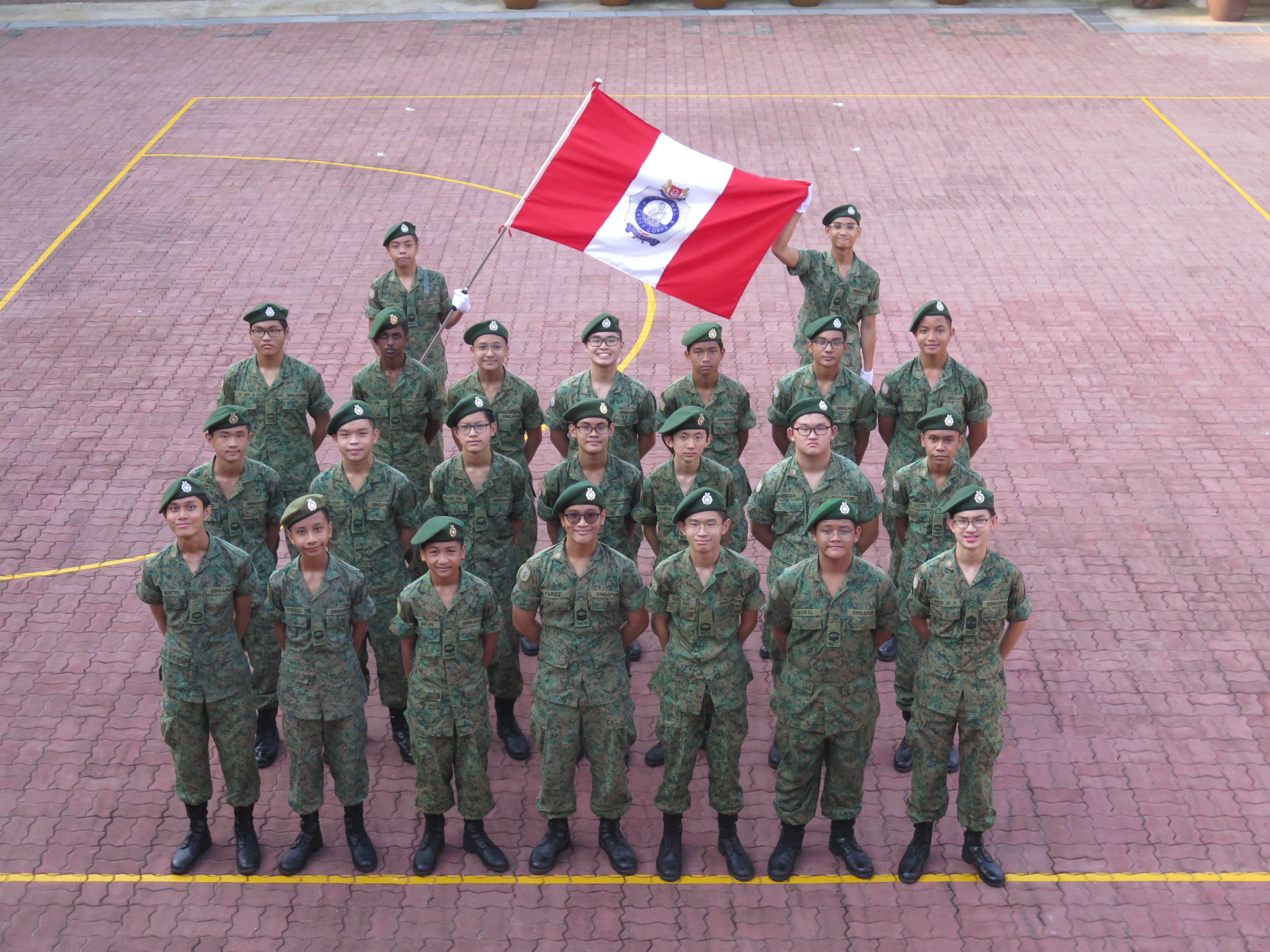 Group of young cadets in camouflage uniforms and green berets posing outdoors, with one holding a red and white flag.